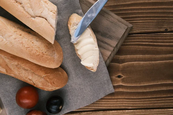 Vista superior baguete francês fatiado fresco com manteiga em um pano de linho com faca e tomate em uma mesa de madeira — Fotografia de Stock