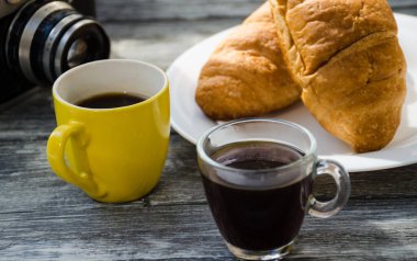 Ahşap arka planda bir fincan kahve ve kruvasanla sakin bir hayat. Eski kamera ve kartpostal bardağın yanında..