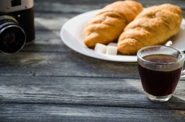 Ahşap arka planda bir fincan kahve ve kruvasanla sakin bir hayat. Eski kamera ve kartpostal bardağın yanında..