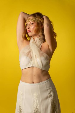 Mujer joven y bella vestida con falda, diadema dorada, y atillo de gasa sobre fondo amarillo, on sesion fotografica en estudio, diosa griega