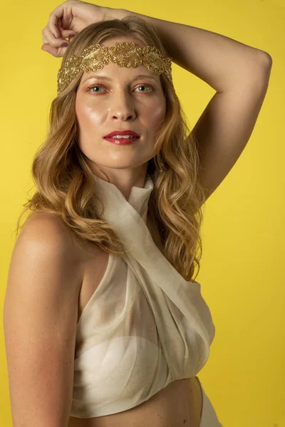 Mujer joven y bella vestida con falda, diadema dorada, y atillo de gasa sobre fondo amarillo, on sesion fotografica en estudio, diosa griega