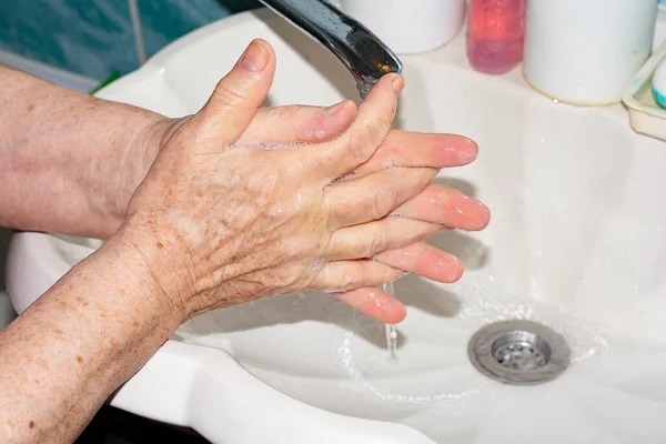 An elderly woman washes her hands under a tap with running clear water. Soapy hands.