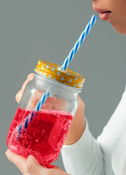 Beyaz bluzlu kadın pipetli bir bardaktan içiyor gri arka planda pembe bir sıvı.