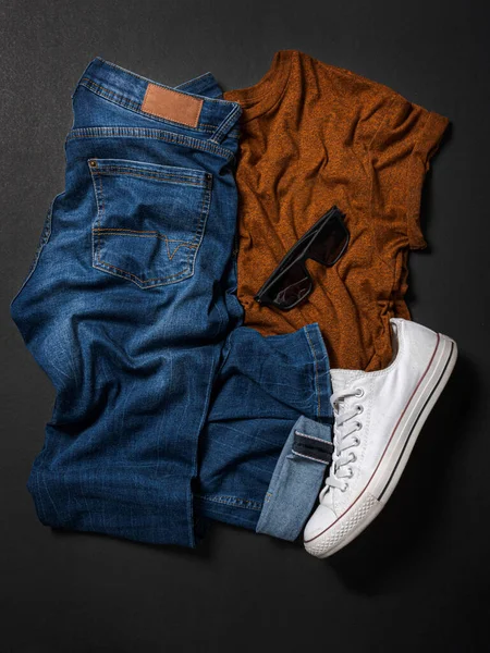 Sarı tişörtlü mavi pantolon buruşuk ve düz zeminde siyah arka planda beyaz ayakkabı.