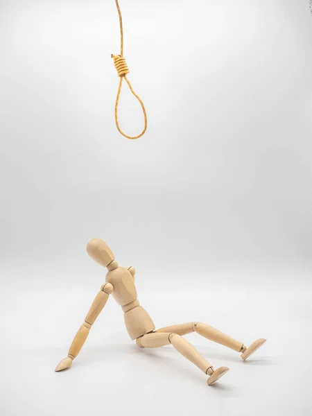 Miniature wooden doll sits under hanging rope and touch gallow noose isolated on white. Concept for thinking in crisis.
