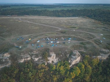 Kayaların üzerinden tırmanma kampı. Açık hava turistik kamp manzarası. Çimenlik alanda çadır ve araçlar var. Uçurumun kenarında patikalar var. Çok renkli turistik evler ve arabalar.