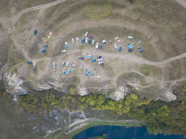 Climbing camp with tents and cars near rocks and cliffs over riverside. Summer day, grass field with pathways and trees. Aerial top down view, drone shot. Russia, Vorgol river.
