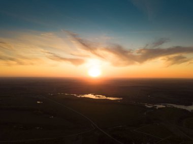 Göllü ve nehirli, karanlık kırsal arazide güneşin doğuşunu gösteren hava manzarası. Ufukta mavi bulutlu gökyüzünde yükselen güneş ve yansıtan suyla birlikte karanlık yüzey.
