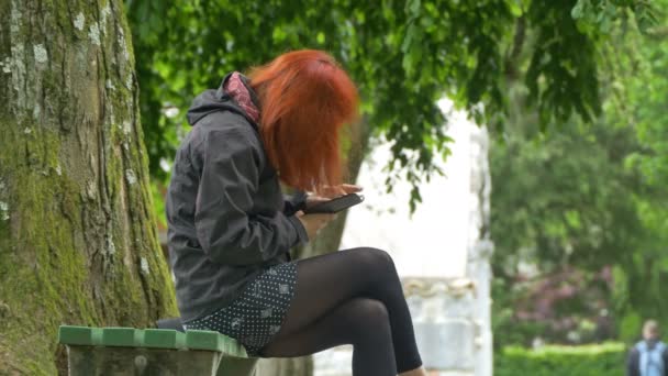赤毛の女の子は ベンチに座って公園でスマートフォブを使用しています — ストック動画