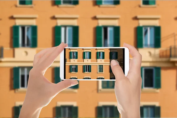 A tourist is making a photo of part of the facade of an orange italian building with windows with green shutters on a mobile phone
