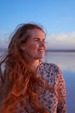 Gün batımında okyanus kıyısında uçan kızıl saçlı güzel, gülümseyen genç bir kadın.
