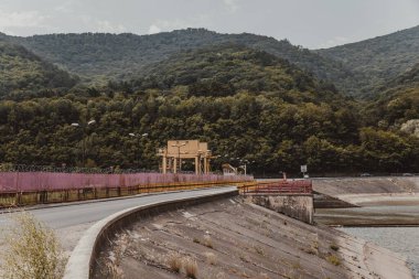 Baraj nehir üzerinde ve bir yol köprüsünde. Aralarından bir nehrin aktığı yüksek dağlar.