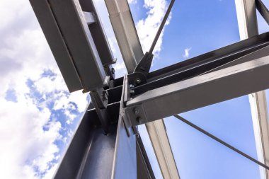 İnşaat halindeki bir binayı destekleyen çelik çerçeve yapısının düşük açılı görüntüsü, arkasında mavi gökyüzü ve beyaz bulutlar var.