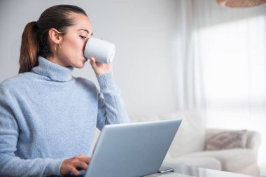 Bir kadın, oturma odasında bilgisayarıyla evden işe koşturuyor.