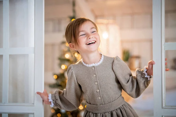 girl in a light interior looks out happily from the door, laughs. A Christmas tree is visible through the glass.