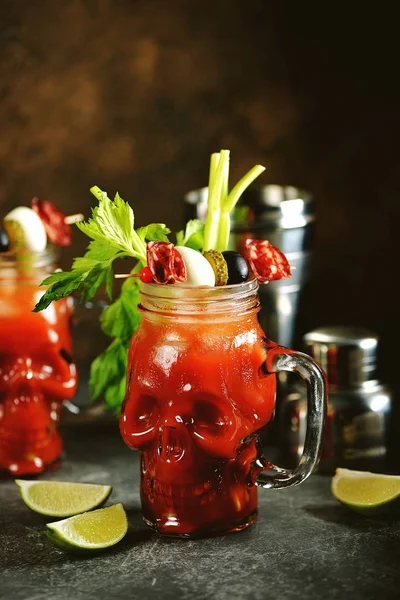 Bloody Mary cocktail in glass skull with celery sticks, pink salt, lime and canapes from canned vegetables. Halloween drink.