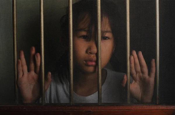 sad asian child standing behind the wire screen window in dark mood.  Unhappy kid alone at home. Upset.