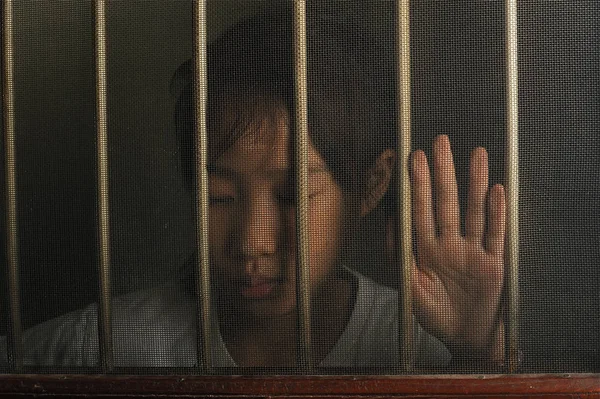 Trauriges asiatisches Kind, das in düsterer Stimmung hinter dem Drahtfenster steht. unglückliches Kind allein zu Hause. Aufruhr im dunklen Raum — Stockbild Trauriges Asiatisches Kind Das Düsterer Stimmung Hinter Dem Drahtfenster Steht — Stockfoto
