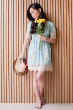 Tam uzunlukta şapka ve buket sarı çiçek ahşap duvara tutan üzgün Asyalı Kız portresi. Kadın hissediyorum yalnız ve hayal kırıklığına moda stil içinde poz.