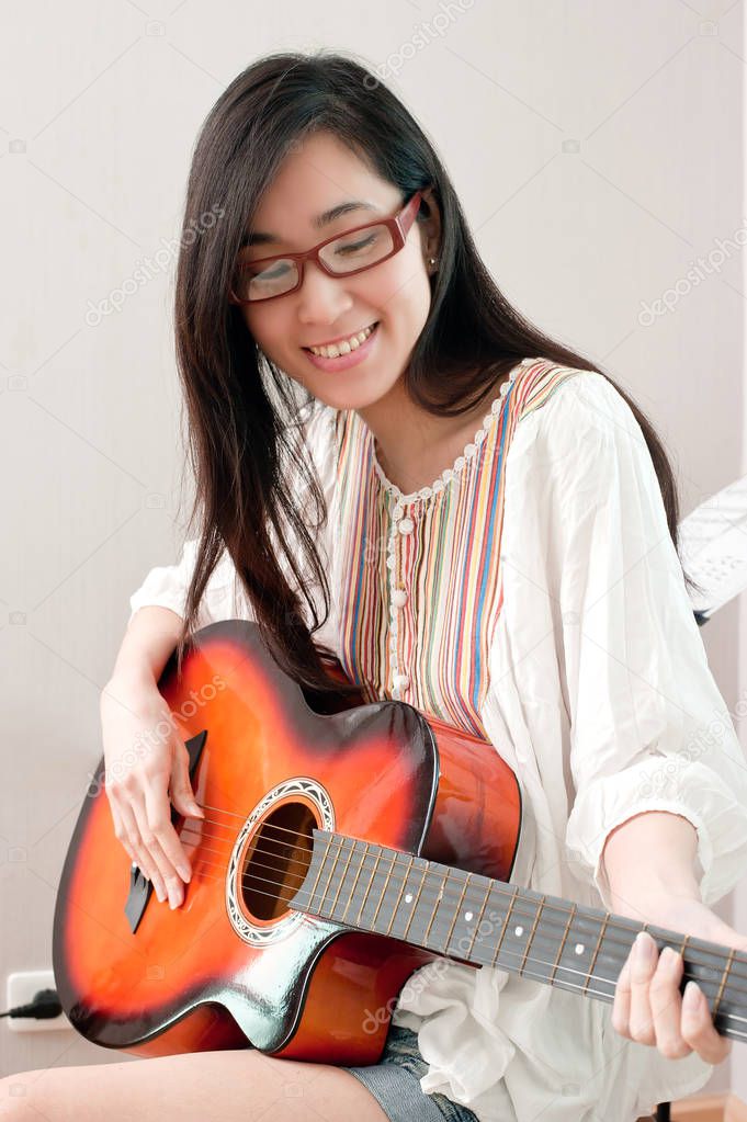 Smiling asian beautiful girl wearing eyeglasses and playing guitar in the music room, leisure concept.