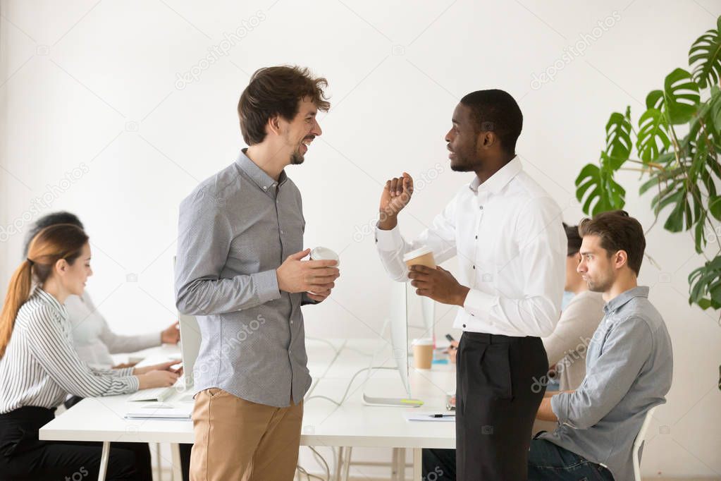 Friendly multiracial young colleagues chatting together in office during coffee break, diverse coworkers talking and laughing at funny joke having good relations and pleasant conversation at work