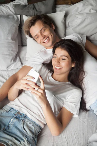 Smiling young couple lying in bed reading news on smartphone — Stock Photo, Image