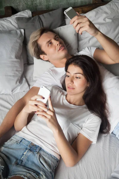 Pasangan milenial yang kecanduan bersantai di tempat tidur menggunakan smartphone — Stok Foto