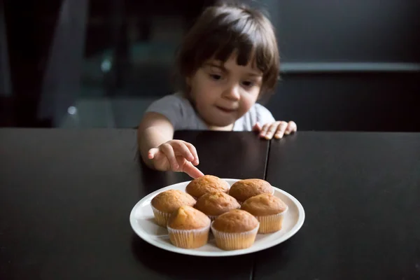 Cute curious little girl reaching muffins on the table — Stock Photo, Image