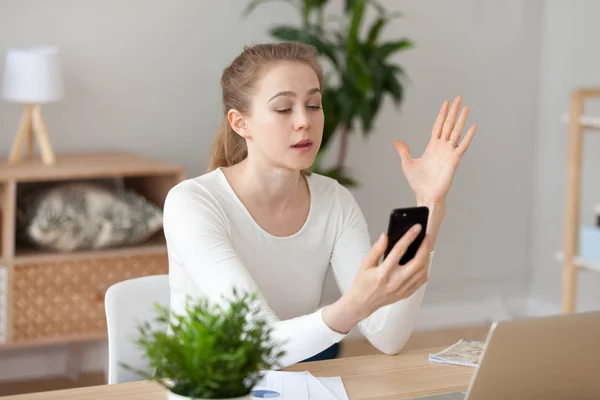 Indignant woman sitting at the desk looking at smartphone screen — Stock Photo, Image