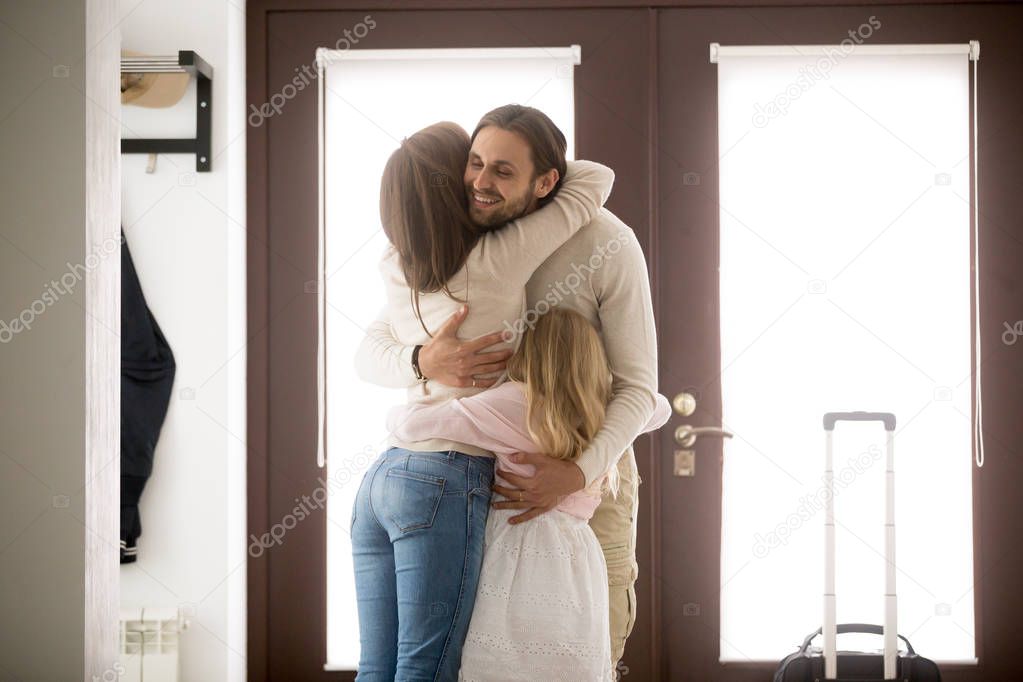 Happy young family in hallway near entrance door at home. Husband hugging wife and little preschool daughter with tenderness and love, with suitcase luggage arrived from business trip, reunion concept