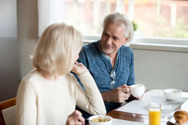 Senior husband look at wife enjoying breakfast in kitchen