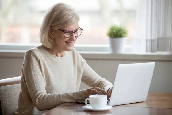 Happy aged woman busy using laptop at home