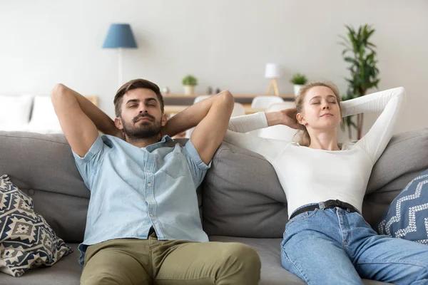 Calm husband and wife relax on couch hands over head