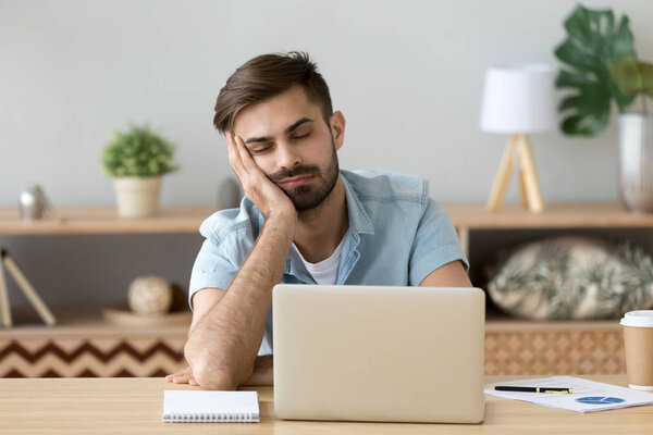 Измученный человек засыпает рядом с ноутбуком на домашнем рабочем месте
