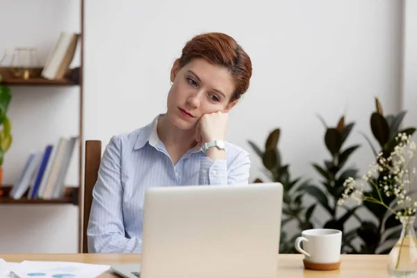 Mujer aburrida sentada en el escritorio mirando la pantalla del portátil —  Fotos de Stock