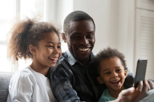 Happy african dad laughing using smartphone with kids at home
