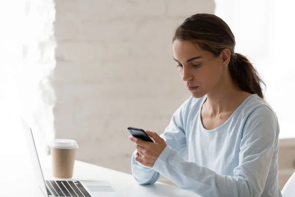 Серйозна зосереджена жінка використовує телефон на робочому місці, читаючи повідомлення — стокове фото