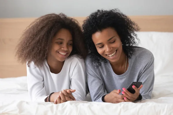 Sorrindo mãe negra e filha relaxar com smartphone na cama — Fotografia de Stock