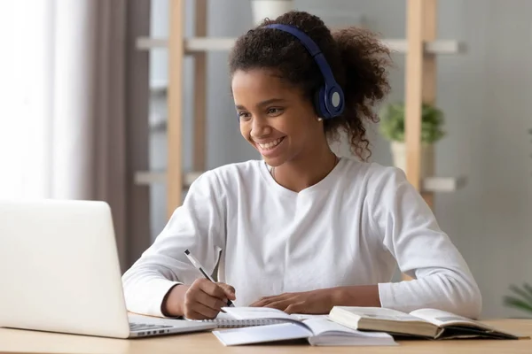 Счастливый черный ученик в наушниках делает домашнее задание — стоковое фото