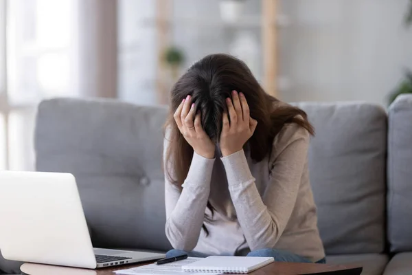 Расстроенная женщина, держащая голову в руках, расстроена, сидя рядом с ноутбуком — стоковое фото