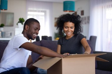 Young black couple open cardboard box unpacking sitting on couch clipart