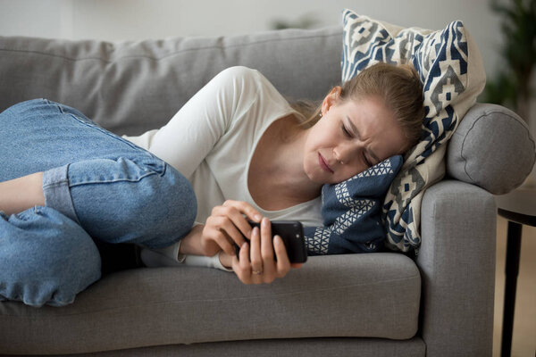 Upset worried young woman crying lying on couch holding phone