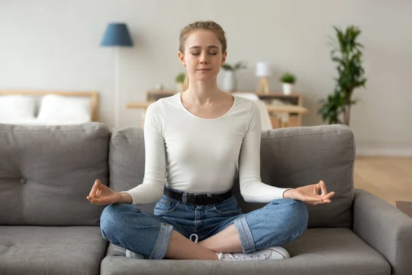 Serene girl sitting on couch doing yoga exercises at home — Stock Photo, Image