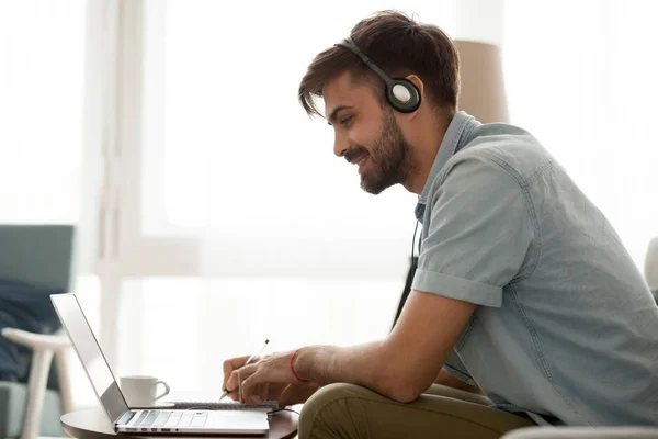 Hombre feliz usando estudio de auriculares en línea en el ordenador portátil haciendo notas —  Fotos de Stock