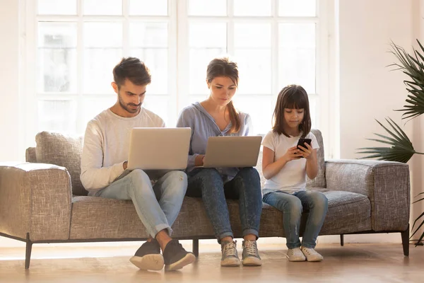 Serious parents with kid daughter sitting on couch using devices