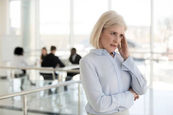 Aged businesswoman suffers from headache standing in office corridor — Stock Photo, Image