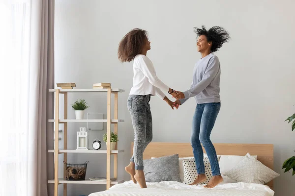 Madre negra e hija adolescente cogidas de la mano saltando en la cama — Foto de Stock