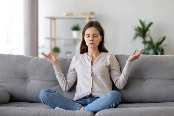 Peaceful young woman sitting in lotus pose on sofa, meditating at home — Stock Photo, Image