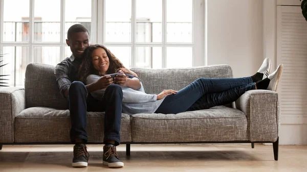 Feliz pareja de raza mixta relajarse en el sofá con el teléfono inteligente —  Fotos de Stock