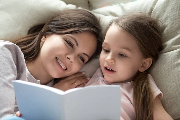 Щаслива мама слухає доньку, яка вчиться читати книгу в ліжку — стокове фото